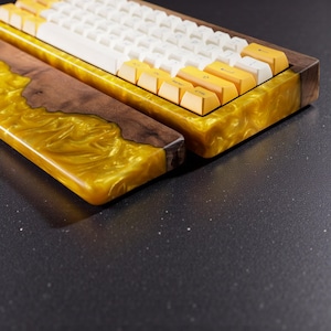 May include: Custom mechanical keyboard with a wooden and resin case. The case features a combination of dark wood and shimmering gold resin. The keyboard has white and yellow keycaps, creating a unique and stylish design.