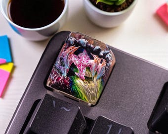 May include: A custom artisan keycap featuring a detailed, colorful dragon design. The keycap is set on a black keyboard, with a cup of coffee and a small succulent plant in the background. The dragon is primarily pink and blue.