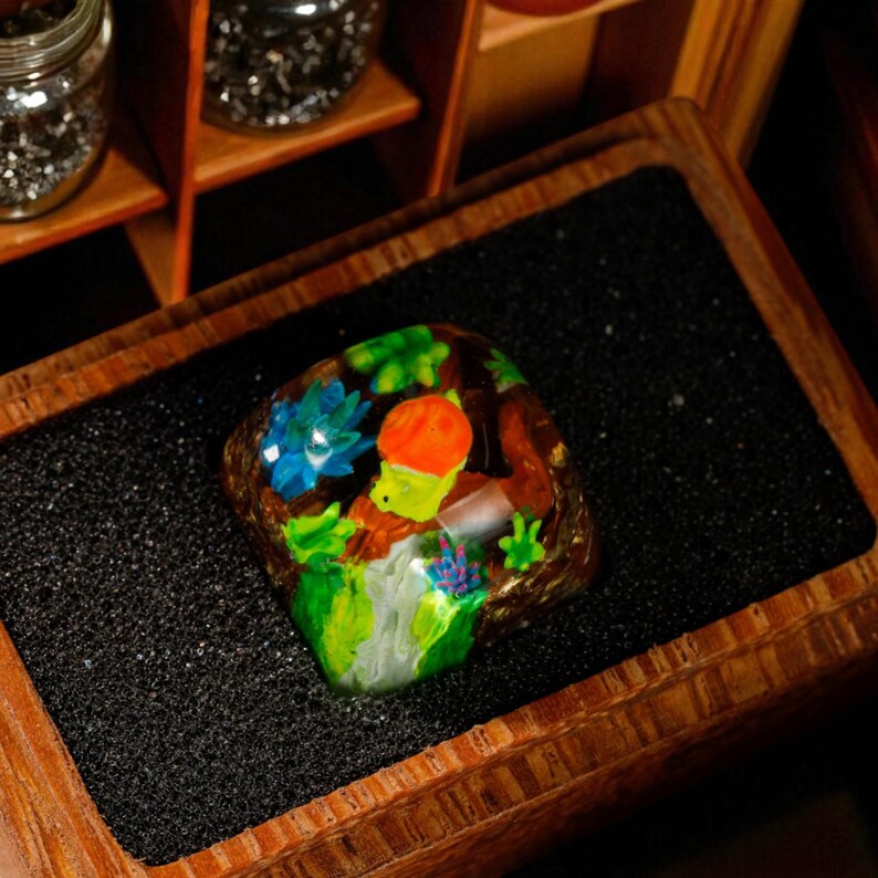 May include: A colorful, resin-cast artisan keycap featuring a whimsical design. The keycap showcases a vibrant scene with a green creature wearing an orange hat, surrounded by blue flowers and green foliage. The keycap is set in a wooden box.