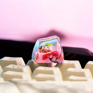 May include: A custom keyboard keycap featuring a miniature landscape scene. The keycap is clear with a colorful interior, including a pink mountain, a red tree, and small figures. The keycap is resting on a keyboard with off-white keys.