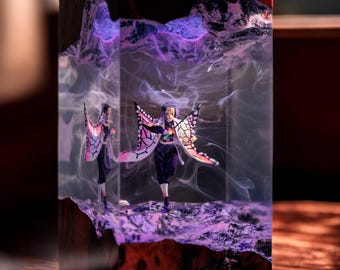 Puede incluir: Una pieza de arte de resina rectangular con una figura con alas de mariposa. La figura está dentro de un bloque de resina transparente con un efecto ahumado púrpura y blanco. La base de madera tiene un tono marrón rojizo.