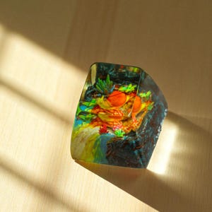 May include: A rectangular, clear resin paperweight with a colorful, miniature landscape inside. The scene includes orange, yellow, green, and blue elements, resembling a vibrant underwater or desert scene. The resin has a dark blue edge.