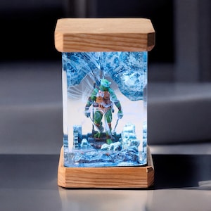 May include: A rectangular resin art piece featuring a Teenage Mutant Ninja Turtle figure. The figure is encased in clear resin with blue and white swirling patterns, set on a wooden base and topped with a wooden lid. The figure is green, with an orange shell and holding two swords.
