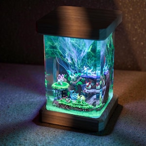 May include: A decorative lamp featuring a miniature forest scene with various figurines. The lamp has a wooden base and top, with a clear, rectangular body. The interior is illuminated with green and blue light, creating a whimsical effect.