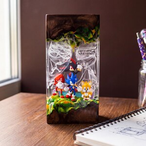 May include: A rectangular resin art piece featuring Sonic the Hedgehog characters. The artwork showcases Sonic, Tails, Knuckles, and Shadow figures set against a backdrop of green, yellow, and brown terrain, with white wisps. The piece is displayed on a wooden surface.