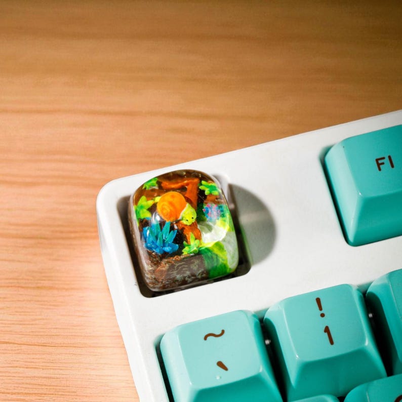 May include: A custom keyboard keycap featuring a miniature underwater scene with orange, green, and blue elements. The keycap is set on a white keyboard with teal keys.