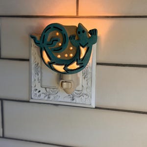 May include: A teal ceramic night light featuring a stylized lizard curled into a circular shape. The design includes cut-out sections that emit a warm glow, and the fixture is mounted on a white, rectangular wall plate with an ornate, embossed pattern.