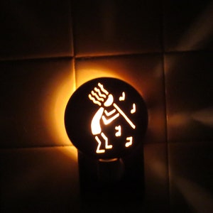 May include: A nightlight with a cutout design of a stick figure holding a spear and surrounded by musical notes. The nightlight is illuminated, casting a warm glow.