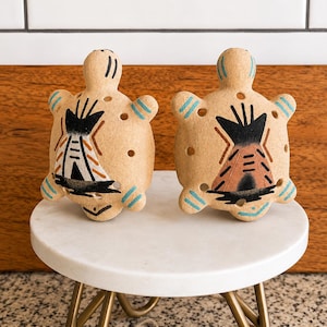 May include: Two tan ceramic turtle-shaped shakers featuring hand-painted black, white, and brown teepee designs on their shells. Each turtle has light blue striped accents on its head and limbs, along with small circular holes on the top surface.