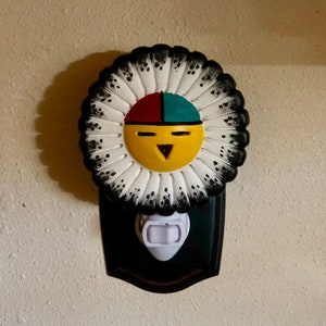 May include: A decorative night light featuring a Native American sun face design. The face is yellow with red and teal accents, surrounded by a white and black feathered headdress. The night light is mounted on a black base.