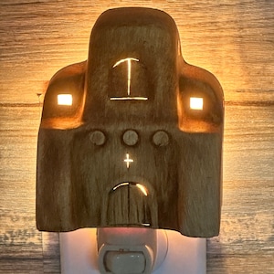 May include: A ceramic night light shaped like a church with a cross on the front and two windows on the sides. The night light is plugged into an outlet and is lit up.