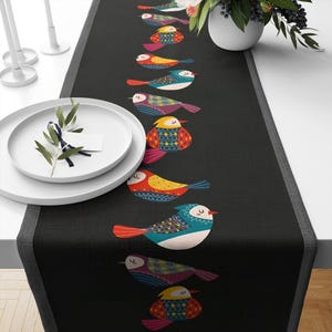 Può includere: Un runner da tavolo nero con un bordo bianco e un motivo ripetitivo di uccelli colorati. Gli uccelli sono stilizzati e hanno motivi geometrici. Il runner è su un tavolo con piatti bianchi.