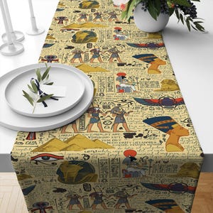 May include: A beige table runner with a repeating pattern of Egyptian hieroglyphs, pyramids, and pharaohs. The pattern is in shades of blue, red, gold, and black. The runner is made of a soft, woven fabric.