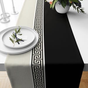 May include: A black and white table runner with a Greek key pattern border. The runner is on a white table with a white plate and a green plant.