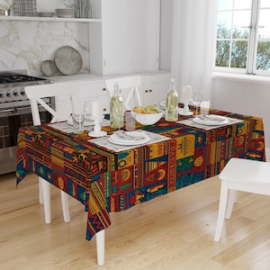 May include: A rectangular tablecloth with a colorful geometric pattern in shades of red, yellow, blue, and green. The tablecloth is set on a white table with place settings, glassware, and a bowl of fruit. The overall theme is dining and home decor.