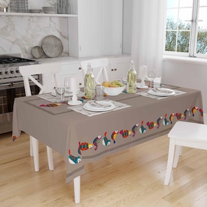 May include: A brown tablecloth with a colorful fish design border. The tablecloth is set on a white dining table with white chairs. There are glasses, plates, and a bowl of fruit on the table.