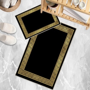 May include: Two black bath mats with a gold Greek key border. One mat is rectangular, and the other is smaller and rectangular. The mats are on a marble-patterned floor. A basket of white towels and a pair of white slippers are visible.