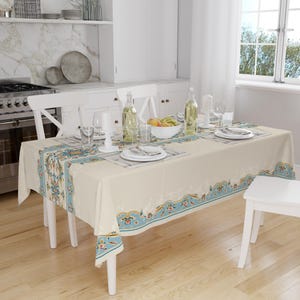 May include: A white tablecloth with a blue and yellow floral border pattern. The tablecloth is set with white plates, glasses, and silverware. There are two bottles of wine and a bowl of fruit on the table.