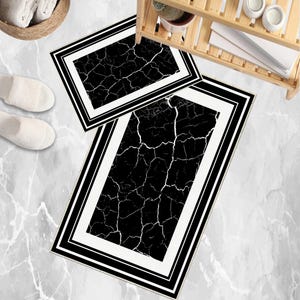 May include: Two rectangular bath mats with a black and white cracked pattern. Each mat has a white border with black trim. The mats are on a marble-effect floor, with white slippers and a wooden shelf in the background.
