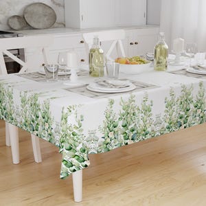 May include: A white rectangular tablecloth with a green and white eucalyptus leaf pattern. The table is set with plates, glasses, and bottles, suggesting a dining setting. The tablecloth is draped over a white table.