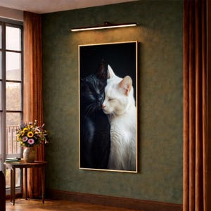 Könnte beinhalten: Ein gerahmter Kunstdruck mit zwei Katzen, einer schwarzen und einer weißen, die sich an den Gesichtern berühren. Die Katzen sind vor einem dunklen Hintergrund platziert, was einen auffälligen Kontrast erzeugt. Das Kunstwerk wird an einer grünen Wand mit warmem Licht darüber ausgestellt.