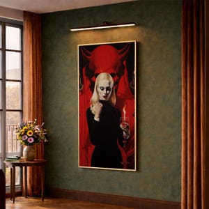 Puede incluir: Una impresión artística enmarcada que presenta a una mujer rubia con un vestido negro, sosteniendo un vaso con una llama, sobre un fondo rojo con una figura de diablo. La obra de arte se exhibe en una pared verde, iluminada por una lámpara.