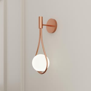 Op de afbeelding: Een koperen wandlamp met een witte bolvormige lamp. De lamp heeft een uniek ontwerp met een gebogen arm en een ronde basis.