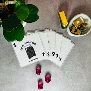 May include: A collection of white Randomizer Cards with black numbers and a suitcase graphic. The cards are arranged with a gold stapler, decorative paper clips, and three pink and red nail designs on a gray surface.