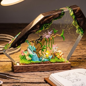 May include: A decorative resin and wood lamp featuring miniature figures of various Eevee evolution characters nestled within a clear, angular resin block. The scene includes a central tree, green foliage, and multiple colorful creature figurines.
