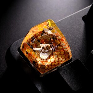 May include: A custom artisan keycap featuring a bee and honeycomb design. The keycap is made of clear resin with a yellow, orange, and brown color scheme. The keycap is designed to fit on a mechanical keyboard.