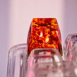 May include: A custom keycap with an orange and white character inside a clear, resin-like material. The keycap has a fiery orange and yellow background, and is set on a clear keyboard.