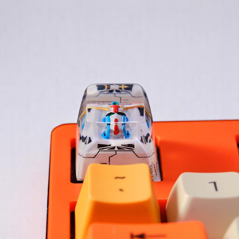 Peut inclure: Un bouton de clavier personnalis&eacute; avec une figurine de petit robot &agrave; l'int&eacute;rieur d'un bo&icirc;tier transparent. Le bouton a un design blanc et gris avec des accents orange et jaune. Le bouton est plac&eacute; sur un clavier orange.