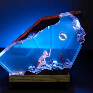 May include: A resin sculpture depicting a scene with a figure reaching for a golden ring. The artwork is encased in a clear, angled structure with a wooden frame and base. The scene includes skulls and figures, all set against a blue backdrop.