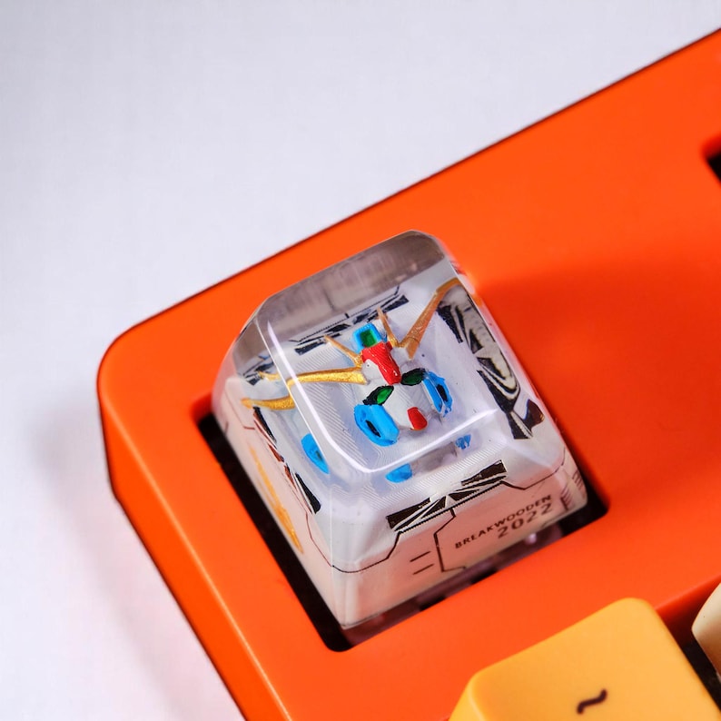 Peut inclure: Un keycap personnalis&eacute; avec un design de robot color&eacute; dans de la r&eacute;sine transparente, pos&eacute; sur un clavier orange. Le keycap pr&eacute;sente des accents dor&eacute;s, bleus, rouges et verts. Le texte "BREAKWOODEN 2022" est visible.