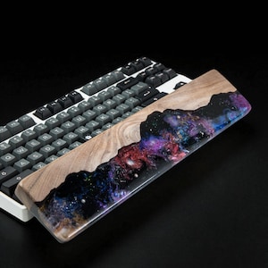 Könnte beinhalten: Eine hölzerne Handballenauflage mit einem Harzeinsatz im Galaxien-Design, vor einer mechanischen Tastatur platziert. Die Handballenauflage hat auf einer Seite eine natürliche Holzmaserung und eine lebendige Mischung aus lila, blauer und roter Harz.