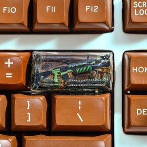 Puede incluir: Primer plano de un teclado con teclas marrones. La barra espaciadora se reemplaza con una tecla de resina transparente que contiene un rifle verde y negro en miniatura. Otras teclas incluyen F10, F11, F12, Inicio y Supr.