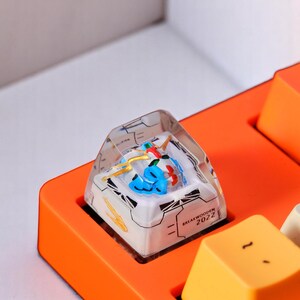 Peut inclure: Un keycap artisanal personnalis&eacute; avec un dessus en r&eacute;sine transparente et un design d&eacute;taill&eacute; avec des &eacute;l&eacute;ments bleus, rouges et jaunes. Le keycap est ins&eacute;r&eacute; dans une base de clavier orange, avec le texte "BREAKWOODEN 2022".
