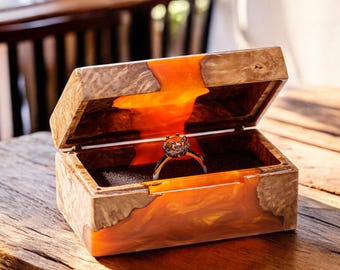 Op de afbeelding: Een houten ringendoos met een transparant oranjehars deksel. De doos is open en er ligt een zilveren ring met een grote diamant in.
