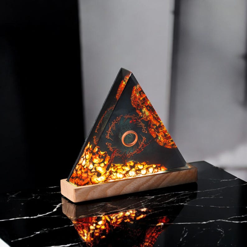 May include: A triangular glass lamp base with a wooden bottom. The glass is filled with a resin-like material with a swirling pattern of orange, yellow, and black. A small, round, copper-colored ring is embedded in the resin.