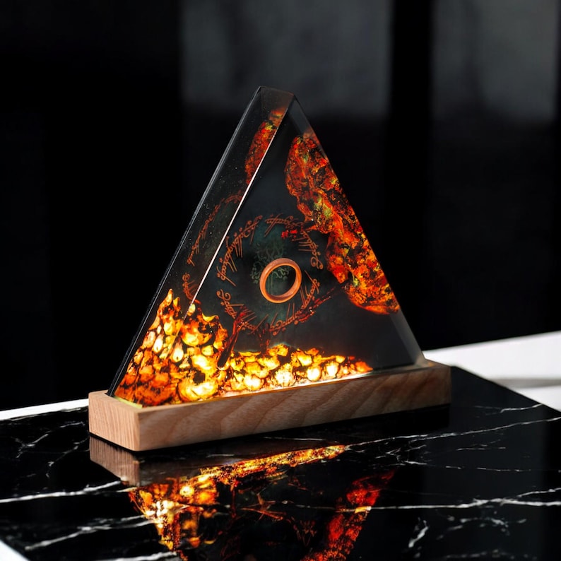 May include: A triangular resin lamp with a glowing orange and yellow base and a dark background featuring a ring and inscription. The lamp sits on a wooden base.