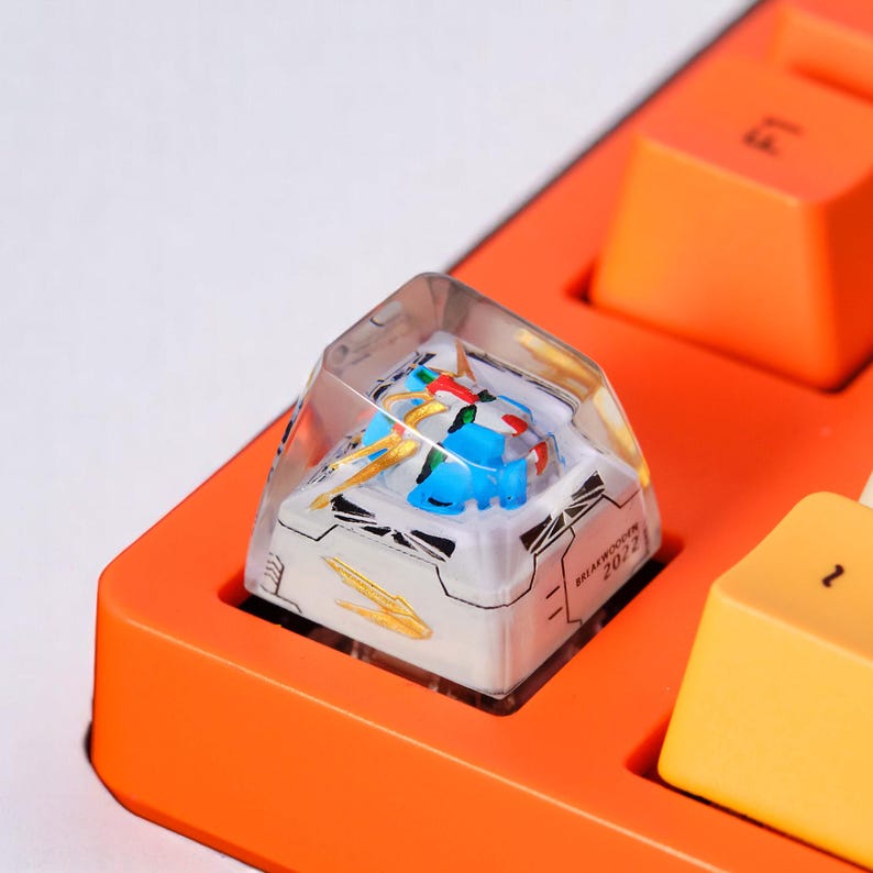 Peut inclure: Un bouton de clavier m&eacute;canique personnalis&eacute; avec un dessus en r&eacute;sine transparente. Le bouton pr&eacute;sente un design de robot bleu et blanc avec des accents dor&eacute;s. Le bouton est ins&eacute;r&eacute; dans une base de clavier orange. Le texte "BREAKWOODEN 2022" est imprim&eacute;.