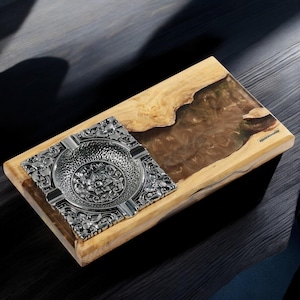 Pode incluir: Um cinzeiro de prata com um design floral intrincado está sobre uma bandeja retangular de madeira com uma incrustação de resina epóxi marrom e verde. A bandeja é etiquetada como "Resinwooden".