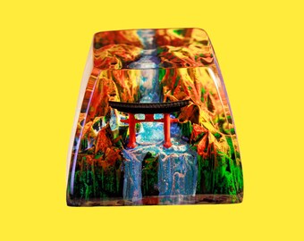 Op de afbeelding: Een transparante hars toetsenkap met een miniatuur rode torii-poort en een blauwe waterval erin. De toetsenkap is omgeven door een kleurrijk landschap met groene, rode en gele accenten.