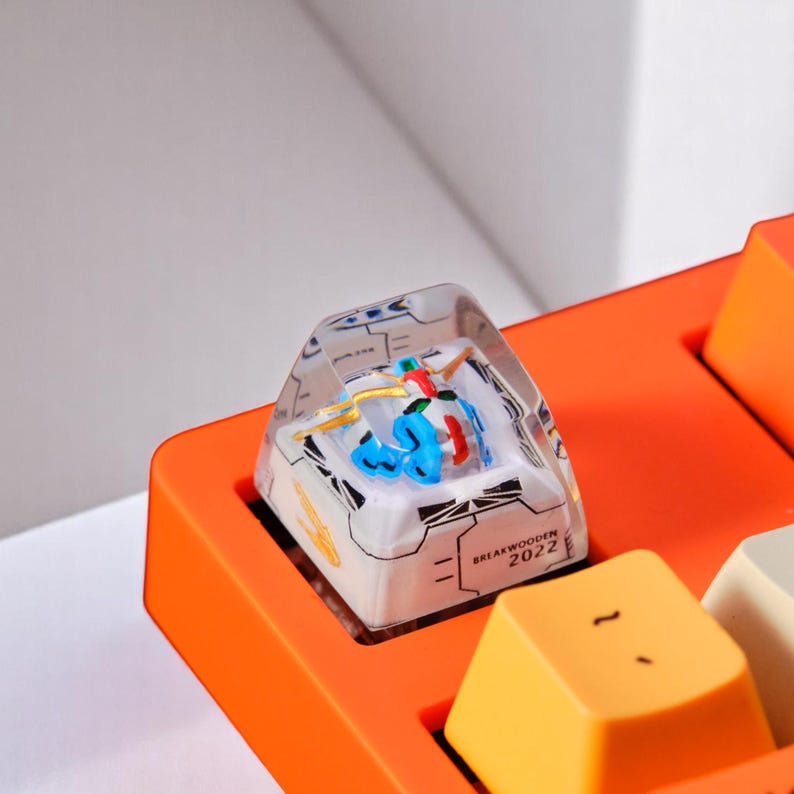 Peut inclure: Gros plan sur une touche de clavier personnalis&eacute;e avec un dessus en r&eacute;sine transparente. La touche pr&eacute;sente un design color&eacute; et d&eacute;taill&eacute; avec des accents bleus, rouges et jaunes. La base est blanche avec des lignes noires et le texte "BREAKWOODEN 2022".