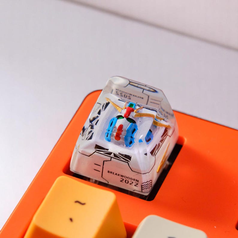 Peut inclure: Une touche de clavier personnalis&eacute;e avec un robot miniature en r&eacute;sine transparente. Le robot a des d&eacute;tails bleus, rouges et blancs. La touche est pos&eacute;e sur une base de clavier orange, avec le texte "BREAKWOODEN 2022".