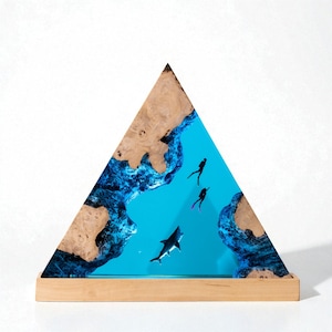 Puede incluir: Una obra de arte triangular de resina que representa una escena submarina con buzos y un tiburón. El marco es de madera natural, con una base de madera. El azul brillante del océano crea un fuerte contraste.