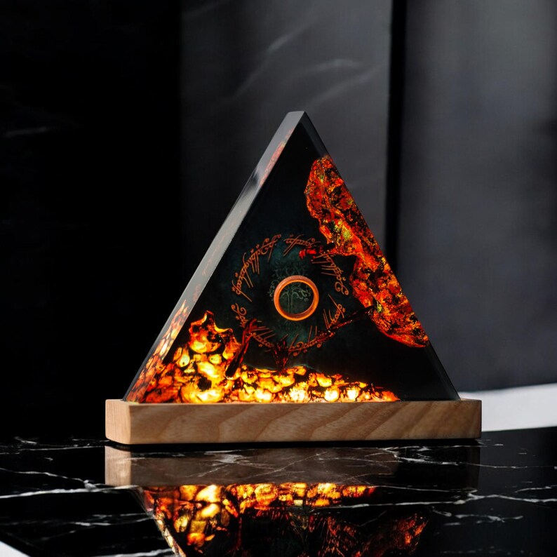 May include: A triangular resin lamp with a glowing orange and green design. The design features a ring and the inscription "One Ring to rule them all, One Ring to find them, One Ring to bring them all and in the darkness bind them." The lamp is sitting on a wooden base.