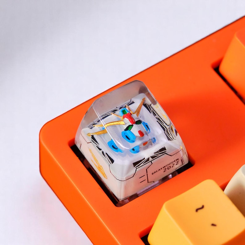 Peut inclure: Un bouton de clavier personnalis&eacute; avec un petit robot &agrave; l'int&eacute;rieur d'une r&eacute;sine transparente. Le robot pr&eacute;sente des accents bleus, rouges et dor&eacute;s, sur un fond blanc avec des motifs g&eacute;om&eacute;triques noirs. Le bouton est ins&eacute;r&eacute; dans une base de clavier orange.