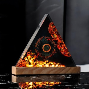 May include: A triangular resin lamp with a wooden base. The resin is clear and contains a depiction of the One Ring from The Lord of the Rings, surrounded by glowing orange and red stones. The inscription "One Ring to rule them all, One Ring to find them, One Ring to bring them all, and in the darkness bind them" is visible on the ring.