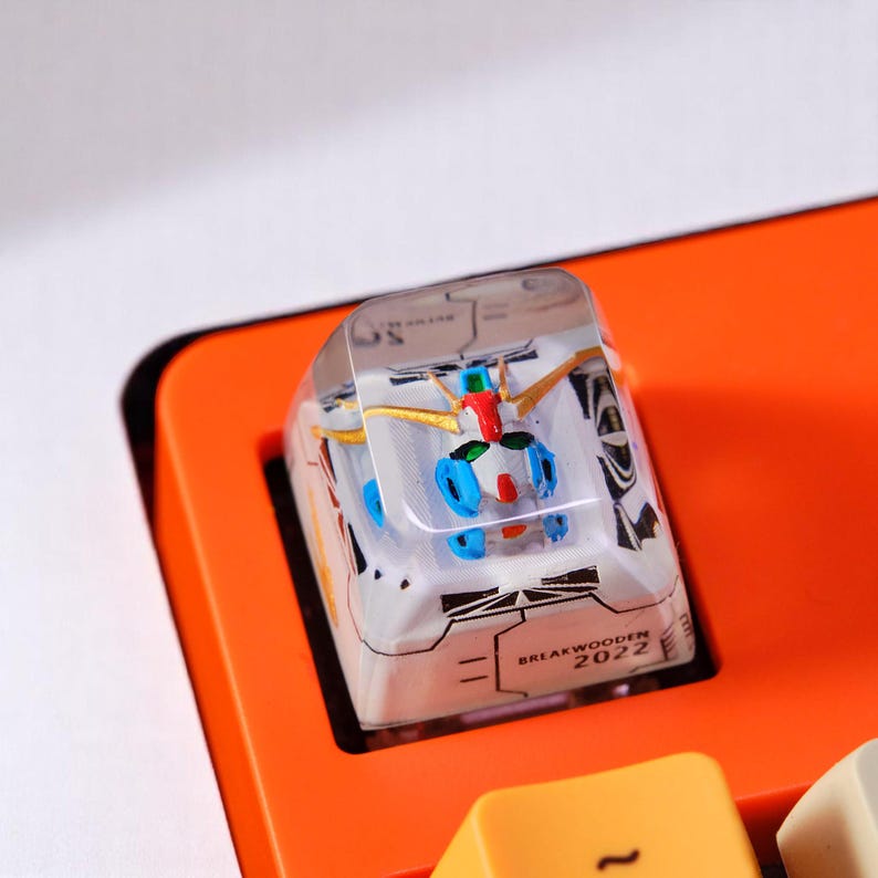 Peut inclure: Un keycap personnalis&eacute; blanc et transparent avec une figurine de robot miniature &agrave; l'int&eacute;rieur. Le robot a des accents bleus, rouges et dor&eacute;s. Le keycap est pos&eacute; sur une base de clavier orange. Le texte "BREAKWOODEN 2022" est visible.